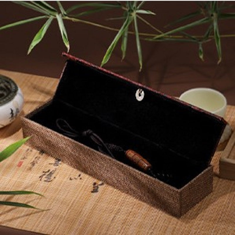 Boîte cadeau zen en lin brun avec un pendentif en bois à l'intérieur, posée sur un tapis en bambou avec des feuilles de bambou en arrière-plan.
