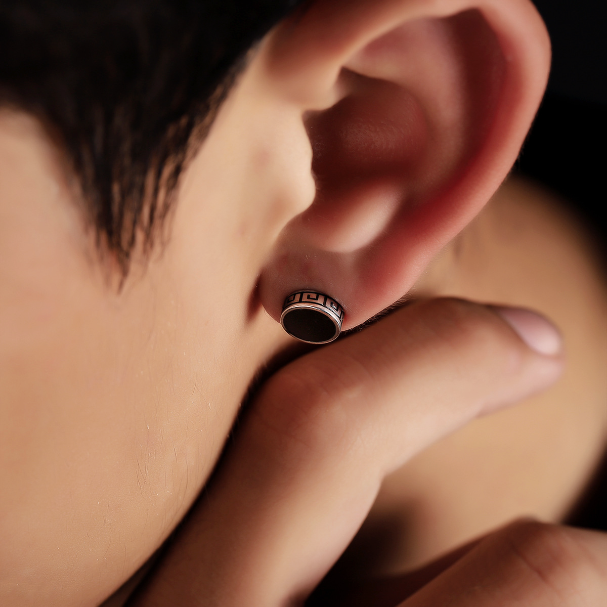 Boucle d'oreille homme argentée et noire avec motif de clé grecque sur le bord, portée à l'oreille.