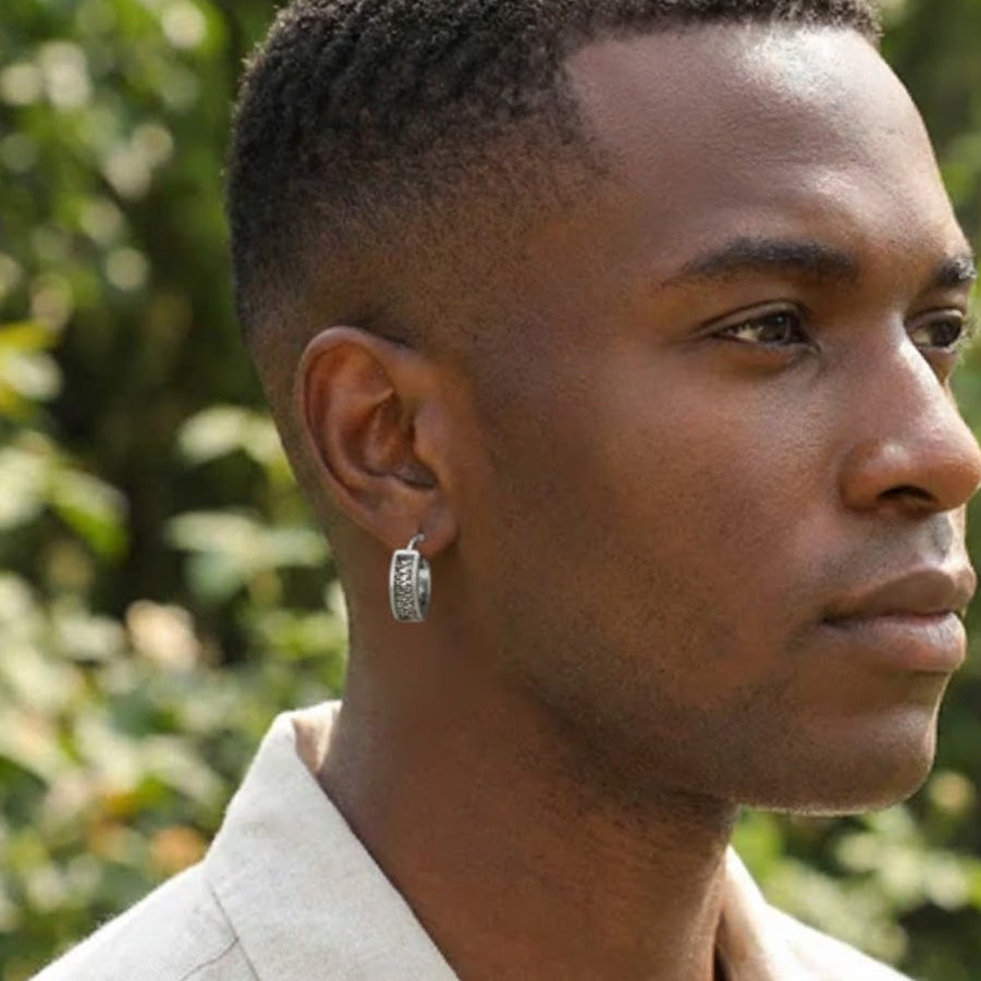 Boucle d'oreille homme argentée style urbain avec motif géométrique gravé, portée par un jeune homme sur fond végétal flou.