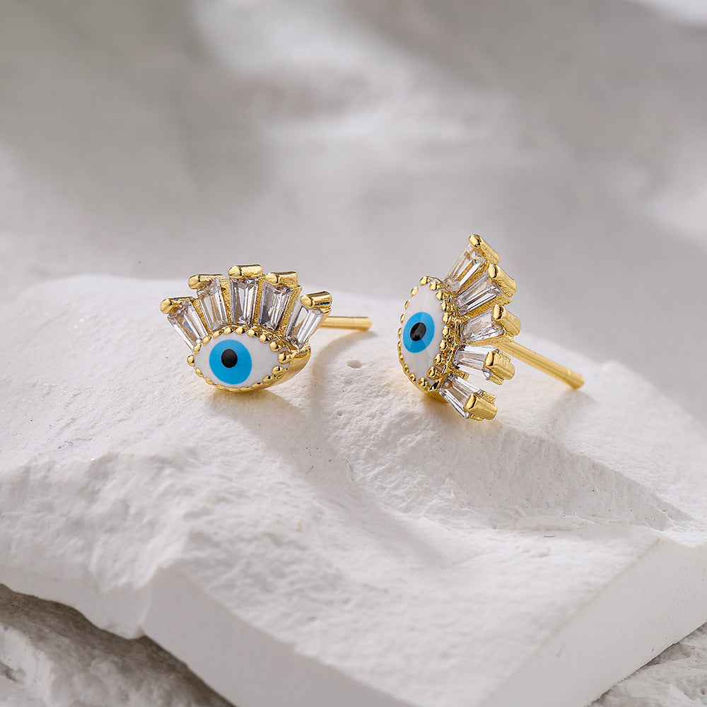 Boucles d’oreilles dorées en forme d'œil bleu stylisé, ornées de cristaux blancs éclatants, posées sur une surface texturée blanche.
