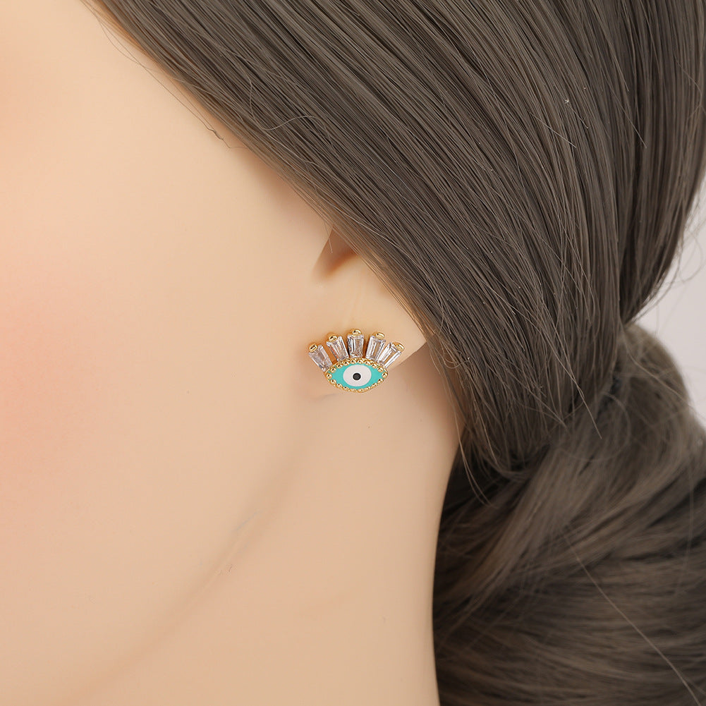 Boucle d’oreille en forme d’œil bleu avec des cristaux blancs sur un fond turquoise, portée sur une oreille humaine