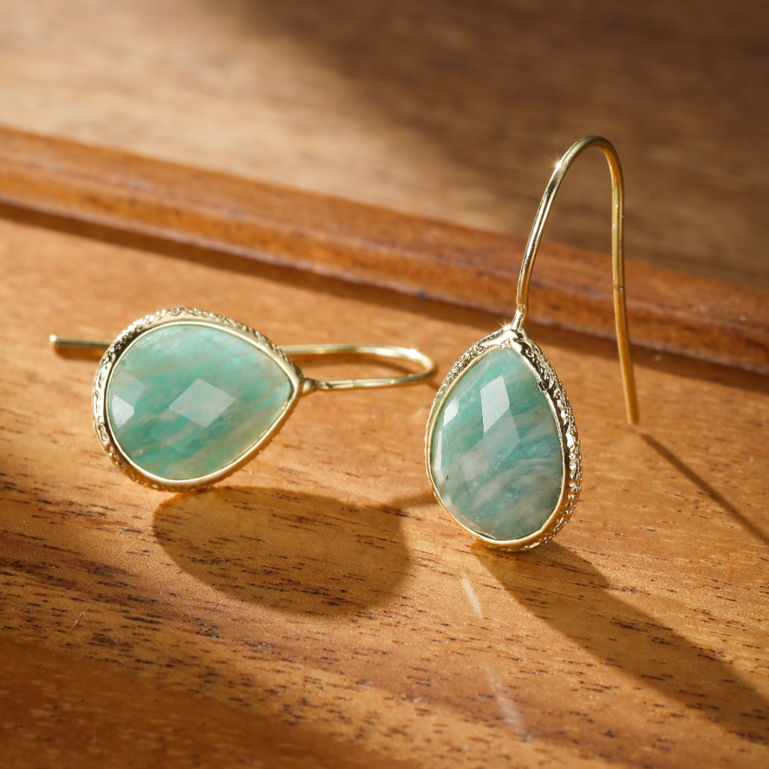 Boucles d'oreilles pendantes en amazonite verte facetée en forme de poire, serties dans un cadre doré texturé, posées sur un fond en bois.