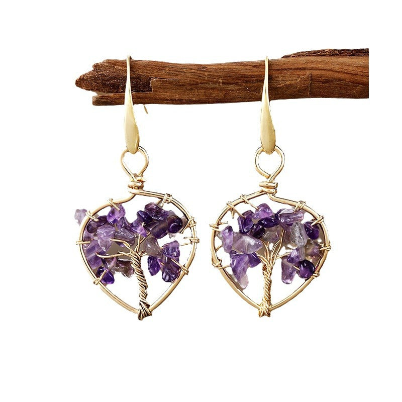 Paire de boucles d'oreilles pendantes en forme d'arbre de vie avec des éclats d'améthyste et des fils de cuivre doré, suspendues à une branche de bois.