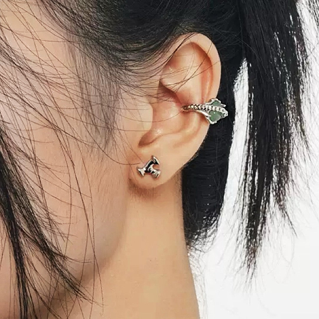 Boucle d'oreille argentée en forme de griffe avec pierre verte, portée sur le cartilage de l'oreille d'une femme, à côté d'un petit clou d'oreille argenté.