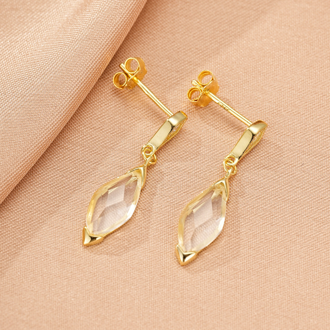 Une paire de boucles d'oreilles pendantes en argent plaqué or 14 carats avec des cristaux de roche taillés en forme de losange, sur un fond rose.