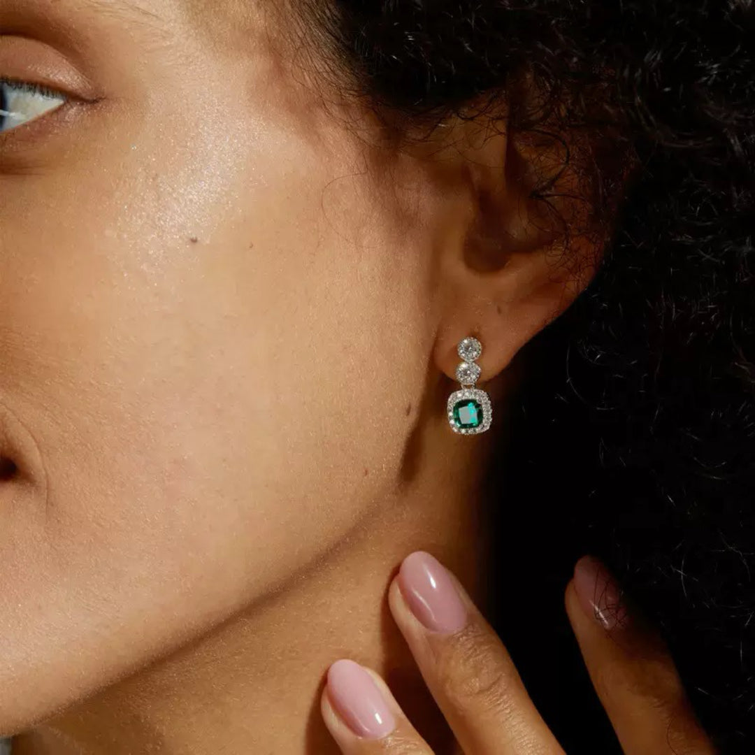 Boucles d'oreilles pendantes en argent plaqué platine ornées d'une émeraude carrée centrale entourée de petits diamants scintillants et de deux diamants ronds au-dessus.