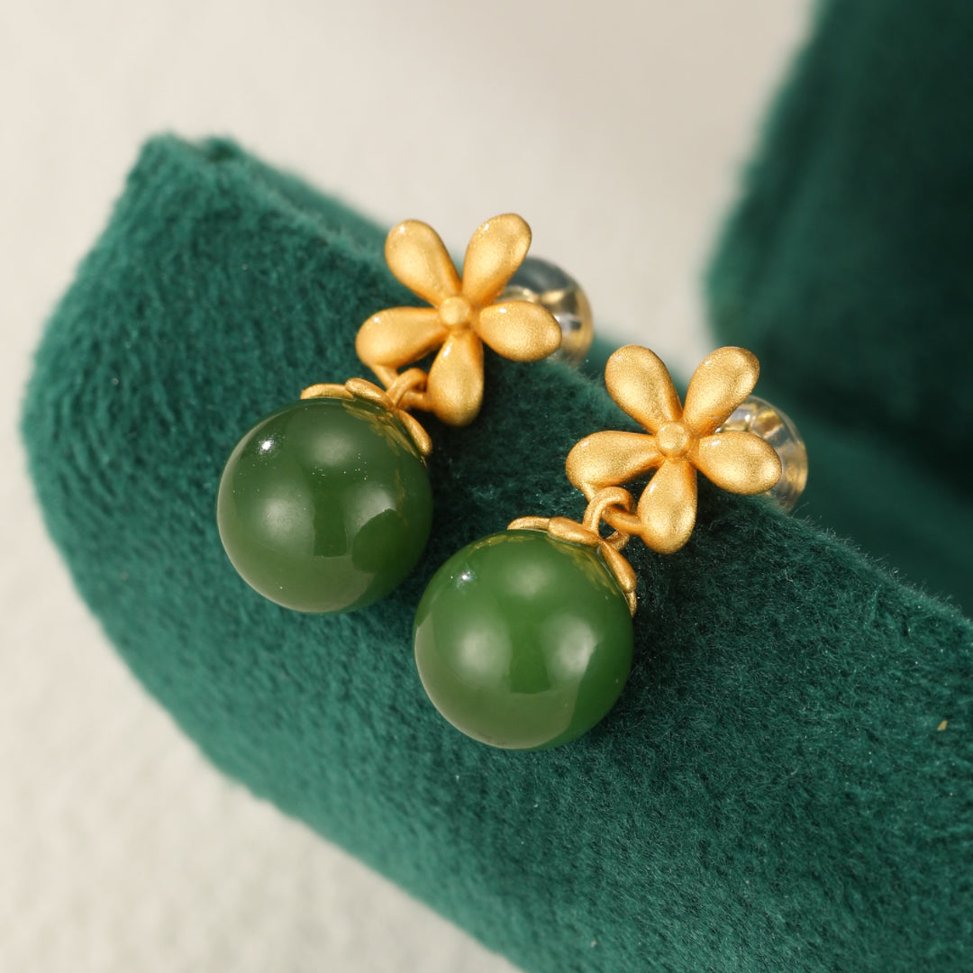 Boucles d'oreilles pendantes en jade vert avec des ornements floraux en argent plaqué or, posées sur un tissu vert foncé.