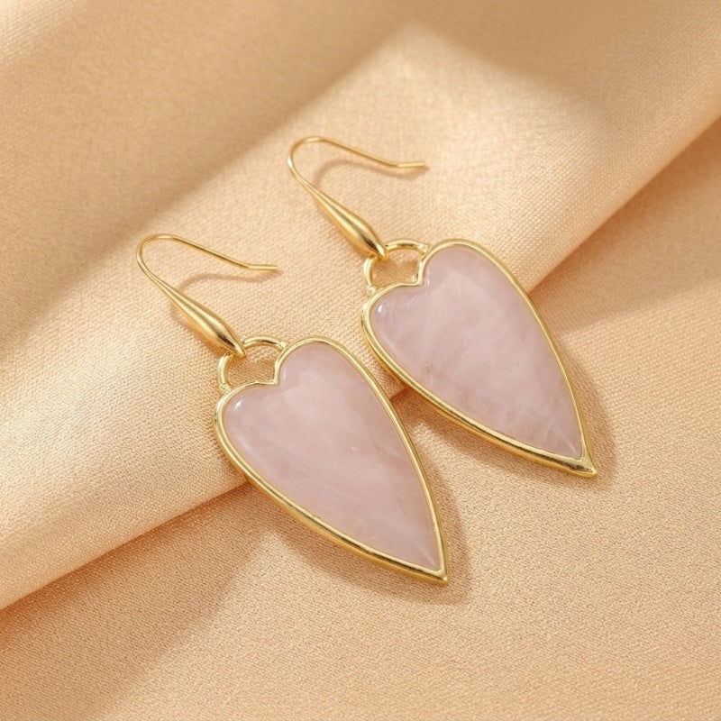 Boucles d'oreilles en forme de cœur en quartz rose avec monture dorée, présentées sur un fond texturé beige.