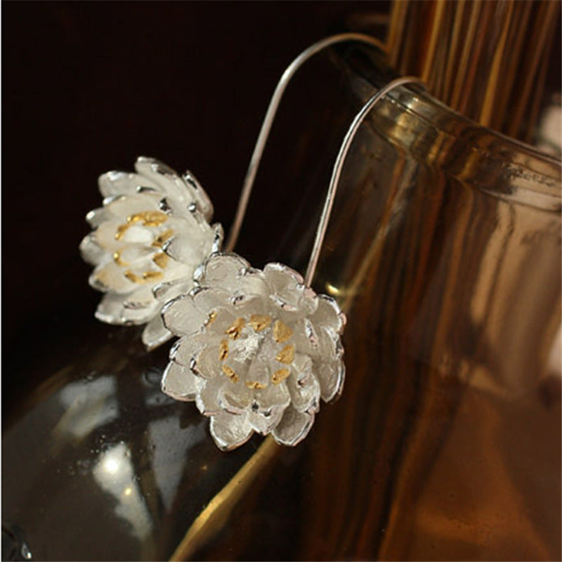 Boucles d’oreilles argentées en forme de fleur de lotus avec des détails dorés au centre, posées sur un fond sombre et réfléchissant.