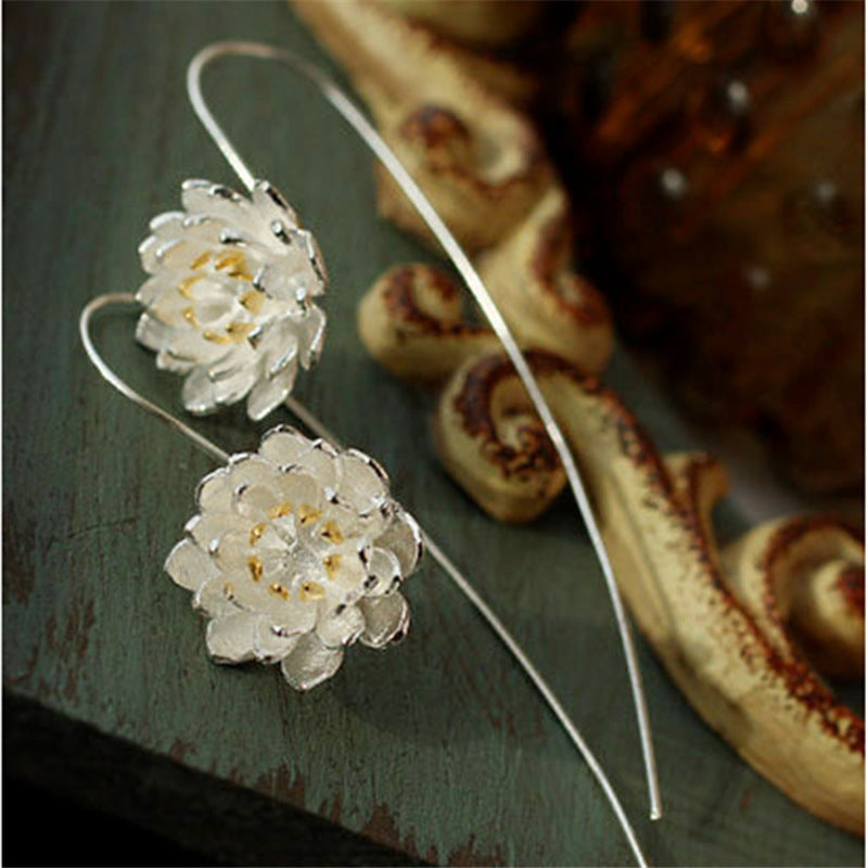 Boucles d'oreilles pendantes en argent avec un design de fleur de lotus détaillée et des accents dorés au centre, sur un fond texturé vert foncé.