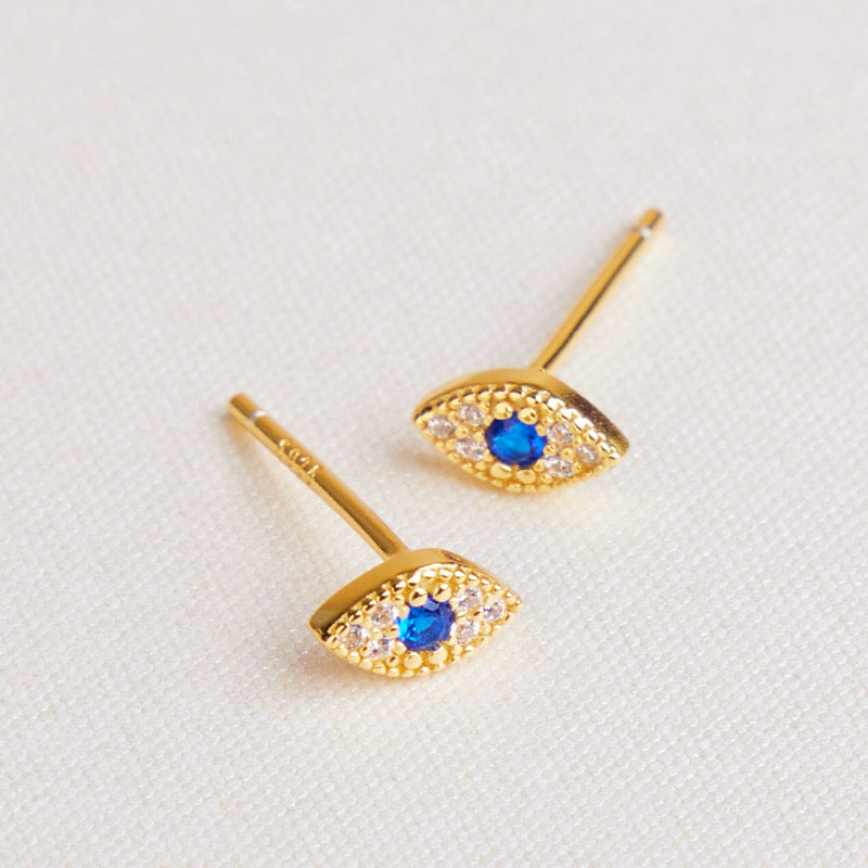 Deux petites boucles d'oreilles en forme d'œil, plaquées or, ornées d'une pierre bleue centrale et de petites pierres blanches scintillantes, sur un fond blanc texturé.