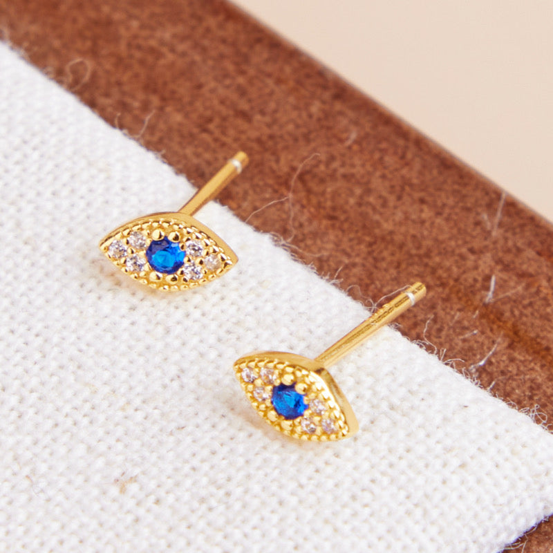Deux petites boucles d'oreilles en forme d'œil, plaquées or, avec une pierre bleue au centre entourée de petites pierres blanches scintillantes.