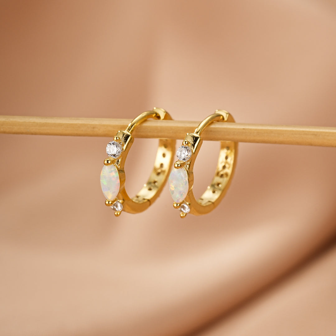 Boucles d'oreilles créoles dorées ornées d'opales marquises et de zirconias cubiques, présentées sur un fond texturé.