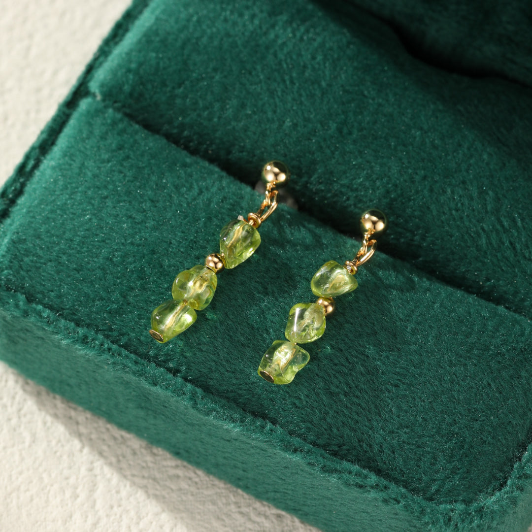 Boucles d’oreilles pendantes en argent plaqué or avec des perles de péridot brut en forme de goutte, posées sur un écrin en velours vert.