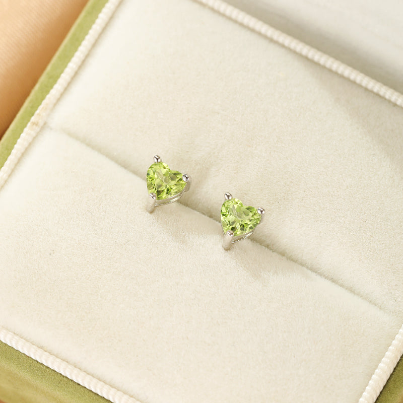 Boucles d'oreilles en argent serties de deux péridots en forme de cœur vert vif, présentées dans une boîte à bijoux verte.