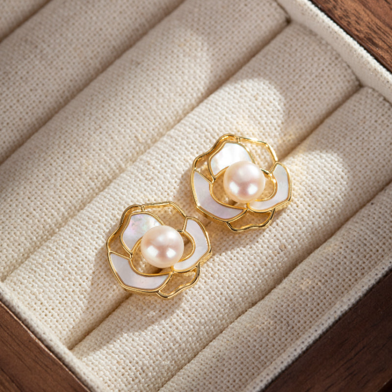 Paire de boucles d'oreilles plaqué or en forme de fleur de camélia avec une perle rose pâle au centre, posées sur un tissu beige dans une boîte à bijoux.