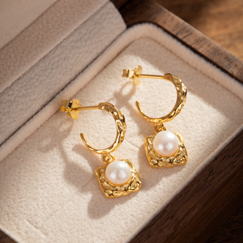 Paire de boucles d'oreilles créoles texturées en argent plaqué or, chacune ornée d'une perle blanche ronde suspendue dans un cadre carré martelé.