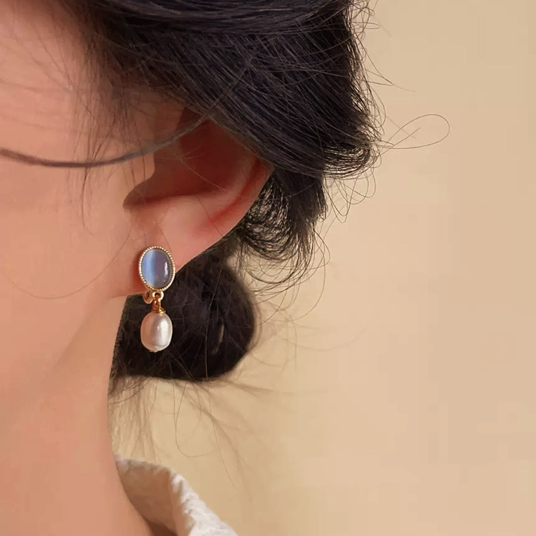 Boucles d'oreilles pendantes avec une perle blanche et une pierre bleue ovale, sur fond beige.