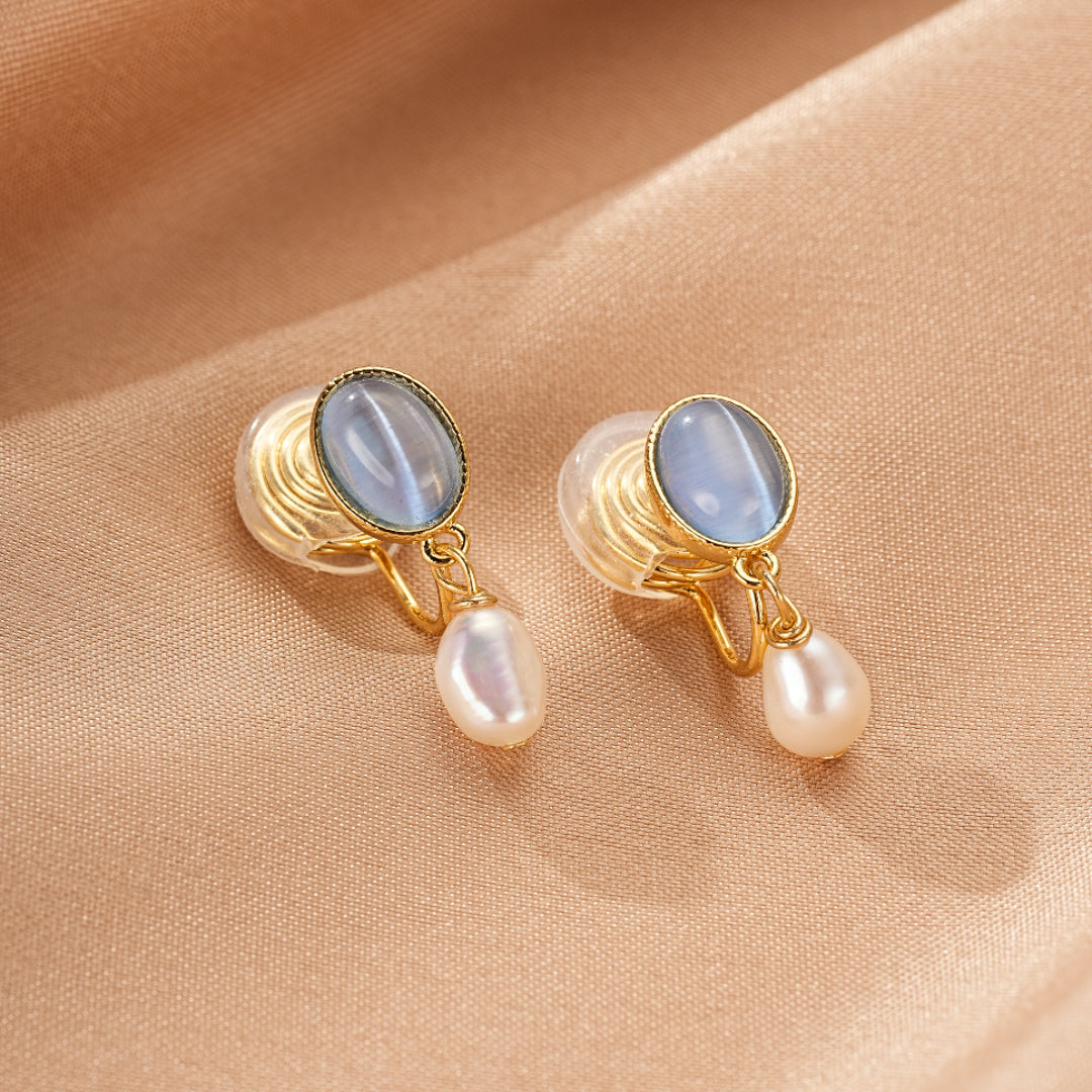 Boucles d'oreilles pendantes avec une perle blanche et une pierre bleue ovale sur fond beige satiné.