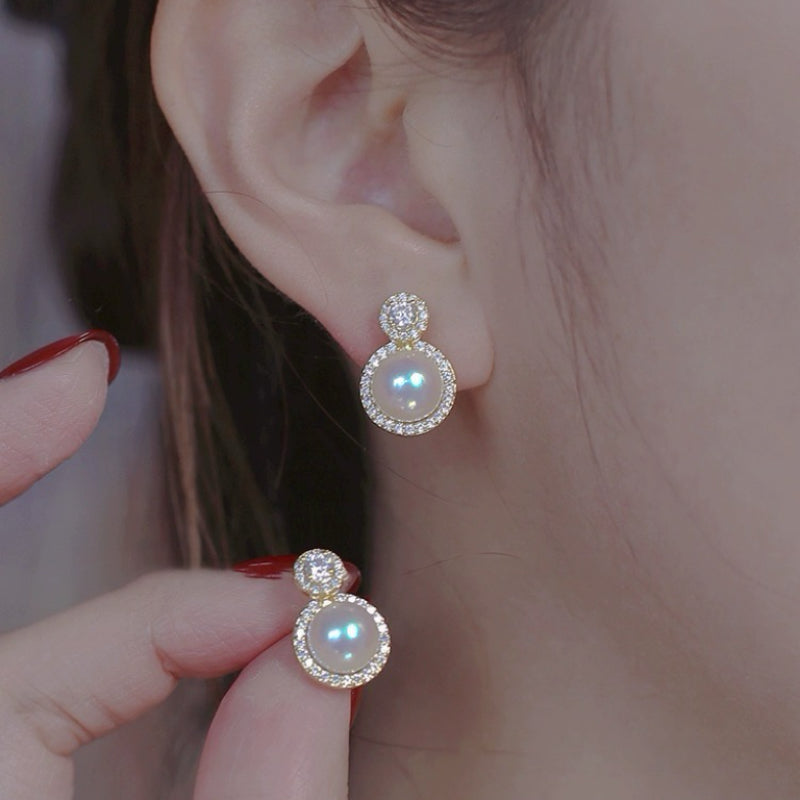Boucles d'oreilles pendantes dorées ornées d'une perle ronde nacrée et d'un cercle de strass brillants, portées à l'oreille d'une femme.