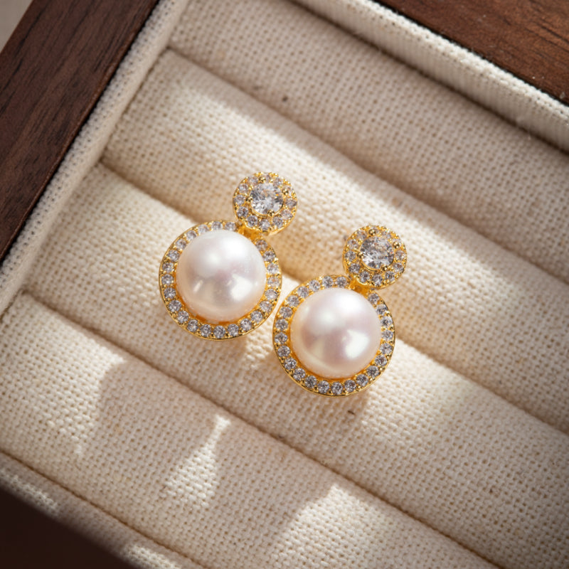 Boucles d'oreilles rondes plaquées or avec une perle blanche au centre, entourée de zirconiums brillants, et un petit zirconium sur le dessus, posées sur un tissu beige.