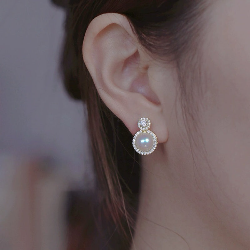 Boucles d'oreilles rondes ornées d'une perle centrale entourée de diamants scintillants, posées sur une oreille.