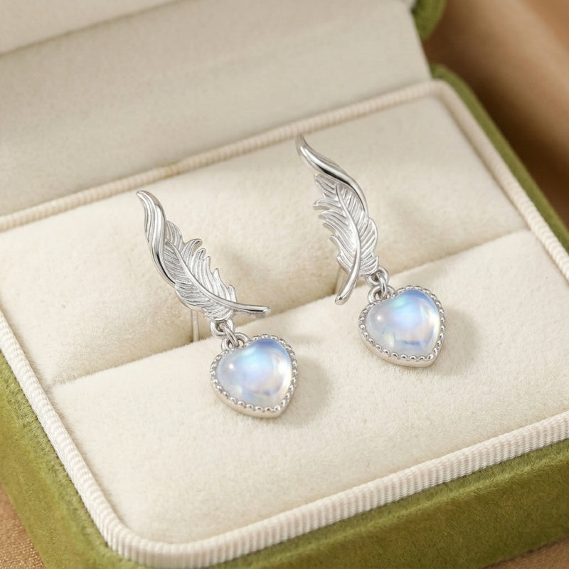 Boucles d'oreilles "Plume Céleste" en argent ornées de pierres de lune bleutées en forme de cœur, suspendues à des motifs de plumes délicats.
