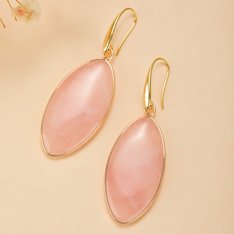 Paire de boucles d'oreilles pendantes en quartz rose ovale avec monture dorée, sur fond beige.