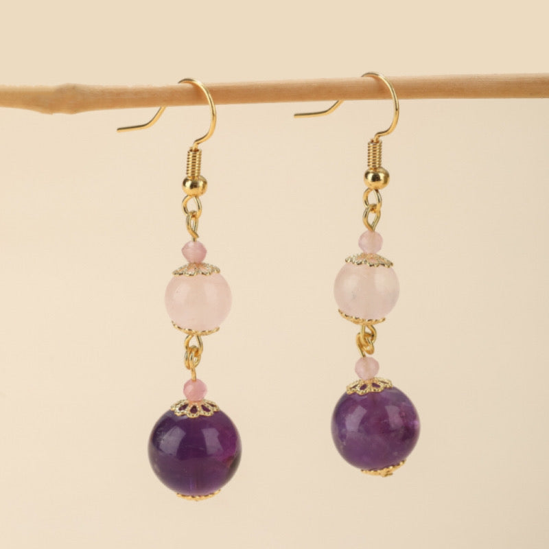 Boucles d'oreilles pendantes en quartz rose et améthyste, avec des perles rondes violettes et roses et des détails dorés, suspendues à un support en bois