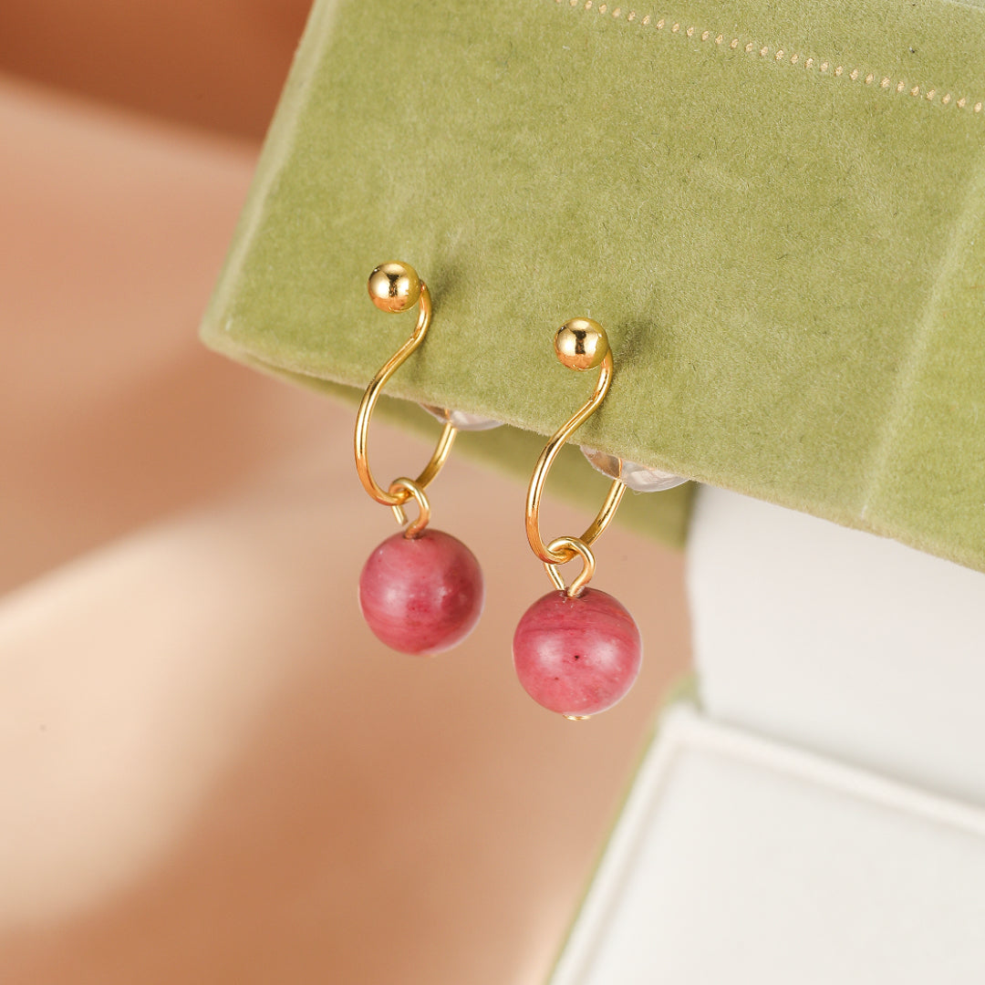 Paire de boucles d'oreilles pendantes en or avec des perles rondes de rhodonite rose, présentées sur un support vert.