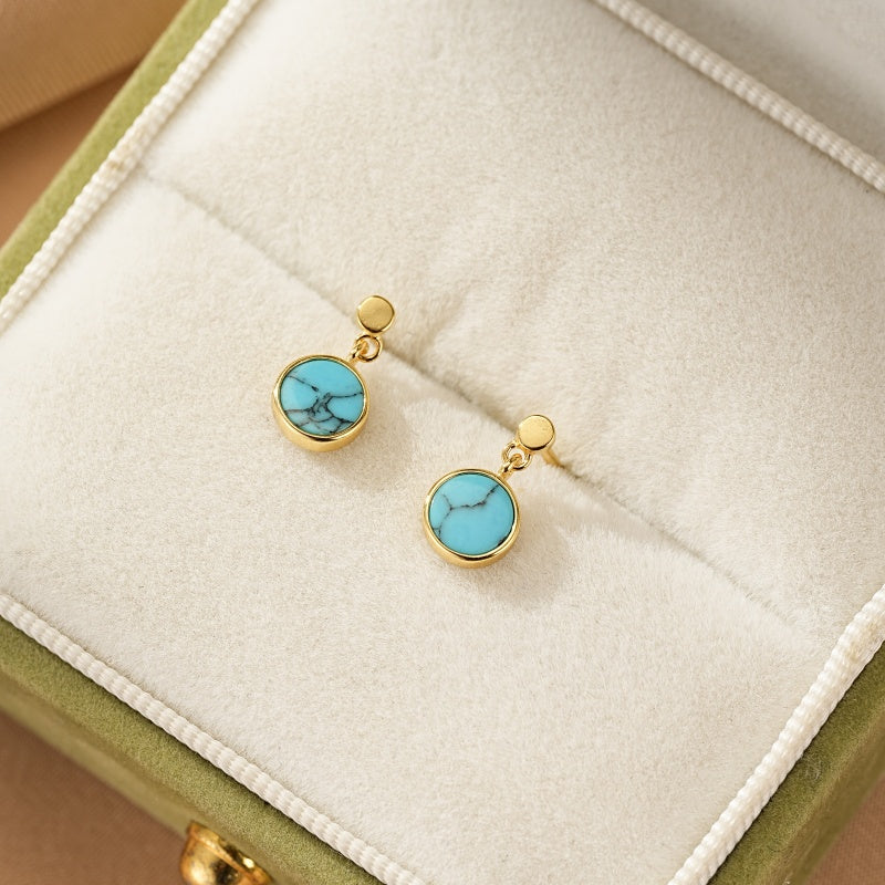 Boucles d'oreilles pendantes Aura du Lagon en argent et plaqué or avec des cabochons ronds en turquoise bleue veinée, présentées dans une boîte à bijoux verte.