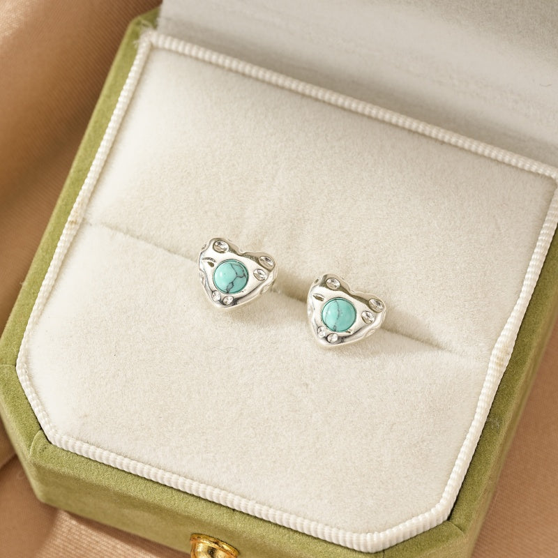 Boucles d'oreilles en argent en forme de cœur avec une turquoise bleue au centre, présentées dans une boîte à bijoux verte.