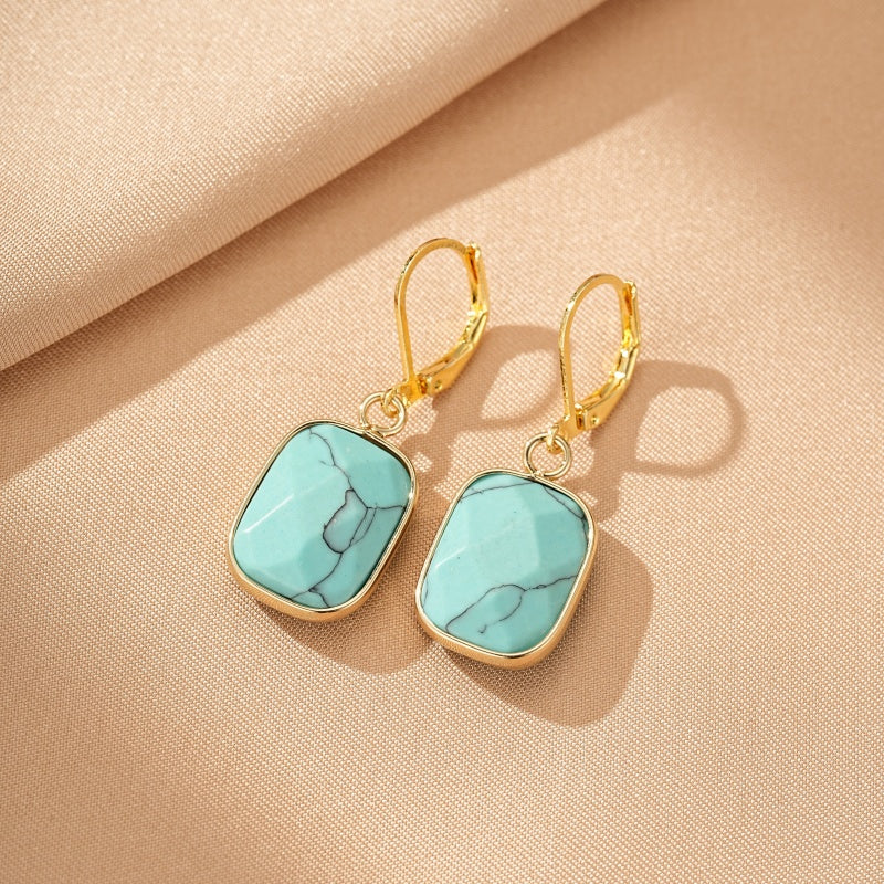 Boucles d'oreilles rectangulaires en turquoise avec monture dorée et fermoir à cliquet, posées sur une toile beige.