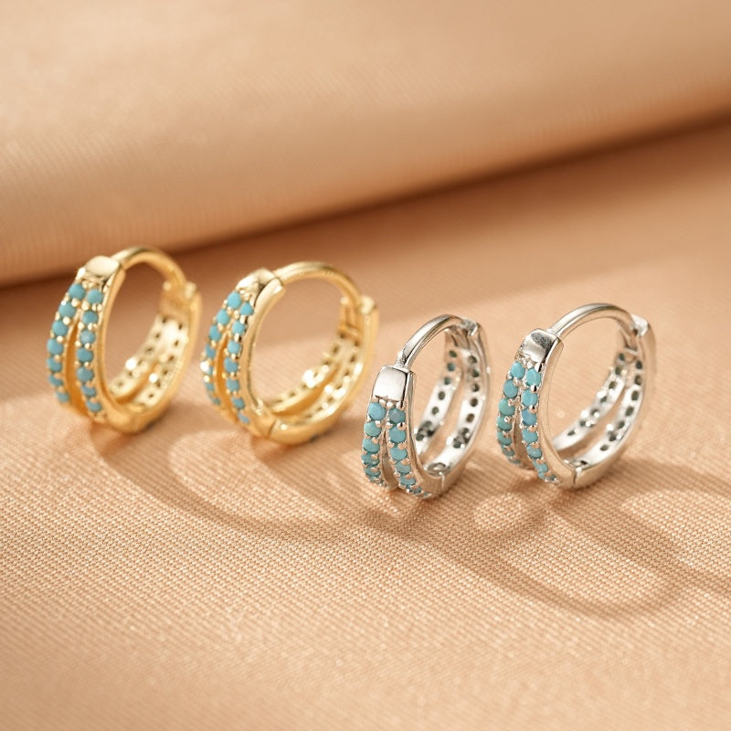 Deux paires de boucles d'oreilles créoles doubles incrustées de petites pierres turquoise, une paire en argent et l'autre en or, sur fond beige.
