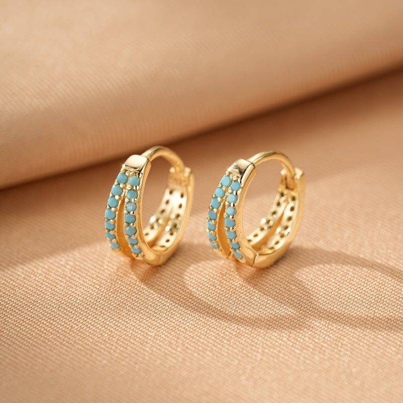Boucles d'oreilles créoles dorées double rangée serties de petites pierres turquoise sur fond beige.