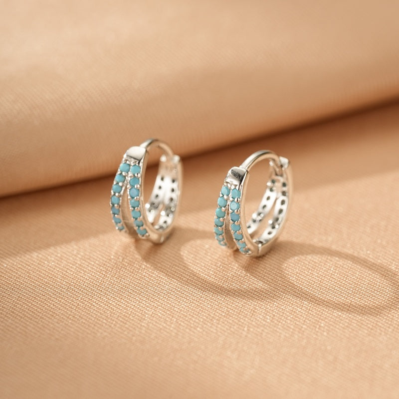 Boucles d'oreilles créoles en argent ornées de doubles rangées de petites pierres turquoise bleues, sur fond beige.