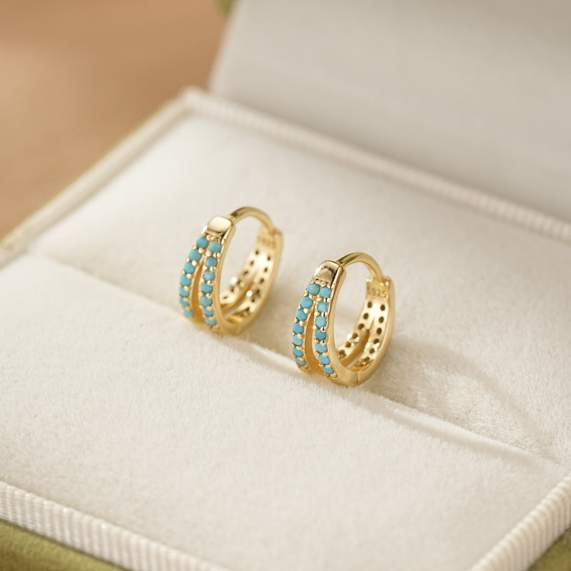 Boucles d'oreilles créoles dorées doubles serties de petites pierres turquoise bleues, présentées dans un écrin vert.