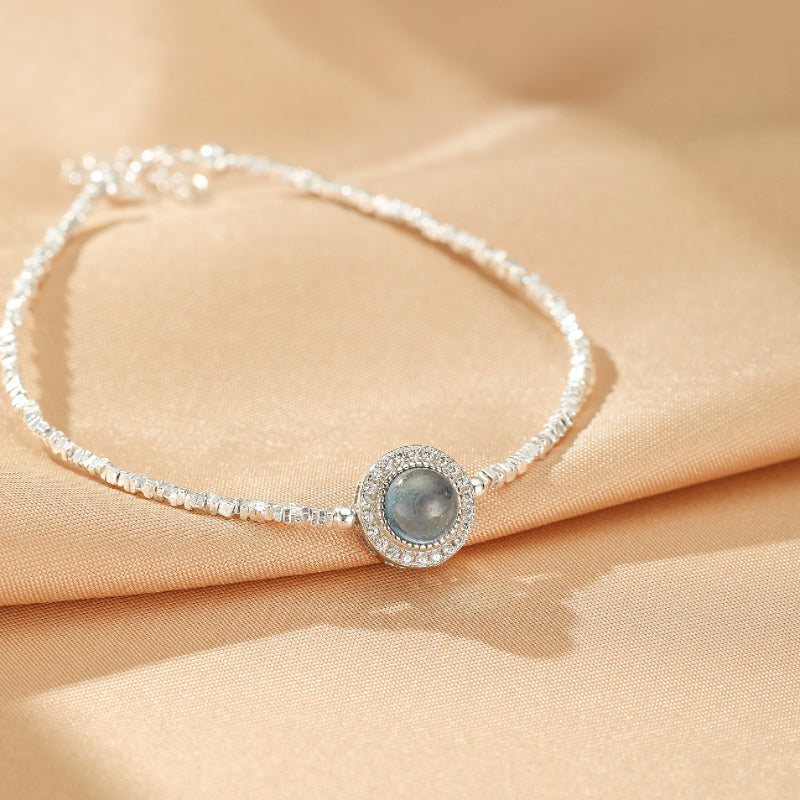 Bracelet fin en argent avec une pierre d'aigue-marine centrale entourée de petits cristaux scintillants, sur un fond de tissu satiné beige.