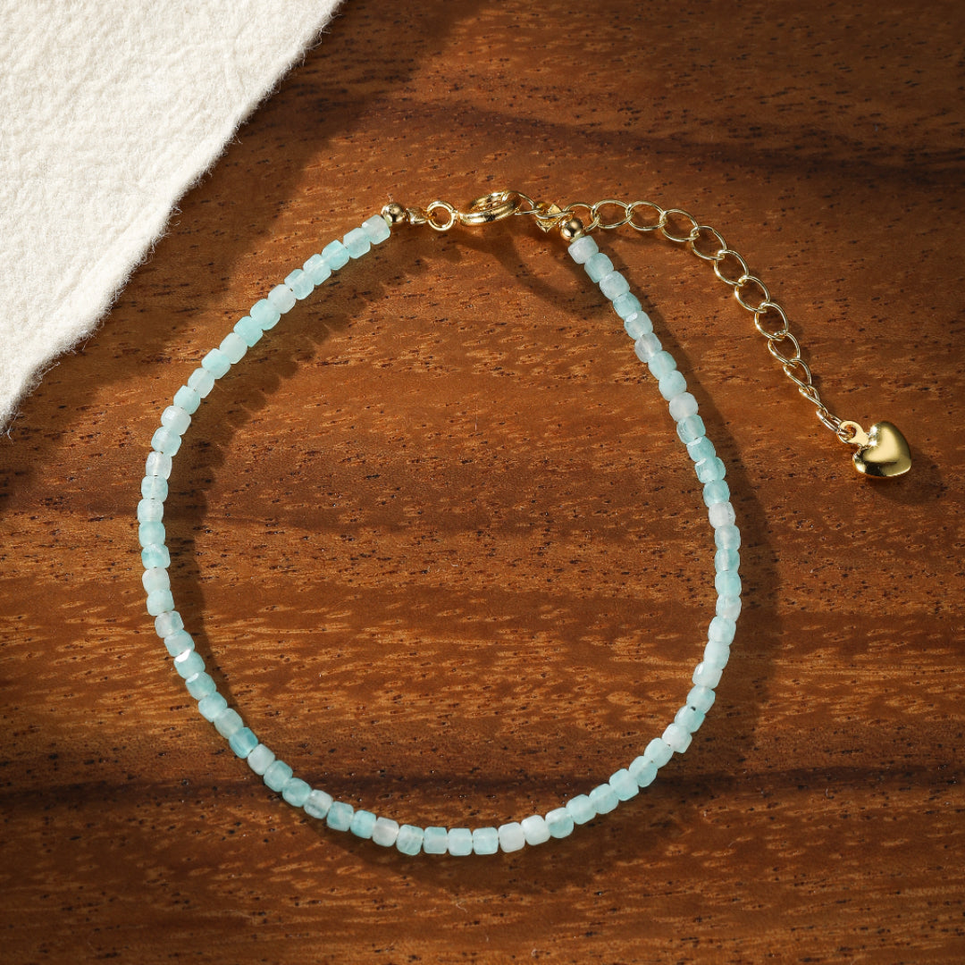Bracelet fin en amazonite bleue clair avec fermoir doré et breloque cœur, posé sur un fond en bois.