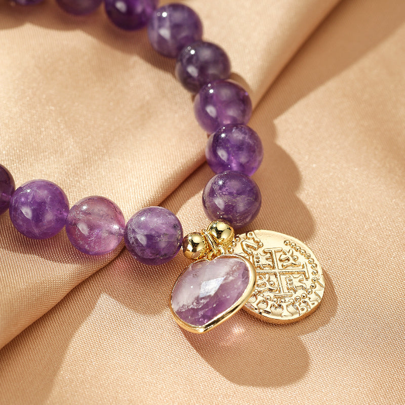 Bracelet "Cœur de Royauté" orné de perles d'améthyste violettes et d'un pendentif en forme de cœur d'améthyste avec une breloque ronde dorée gravée.