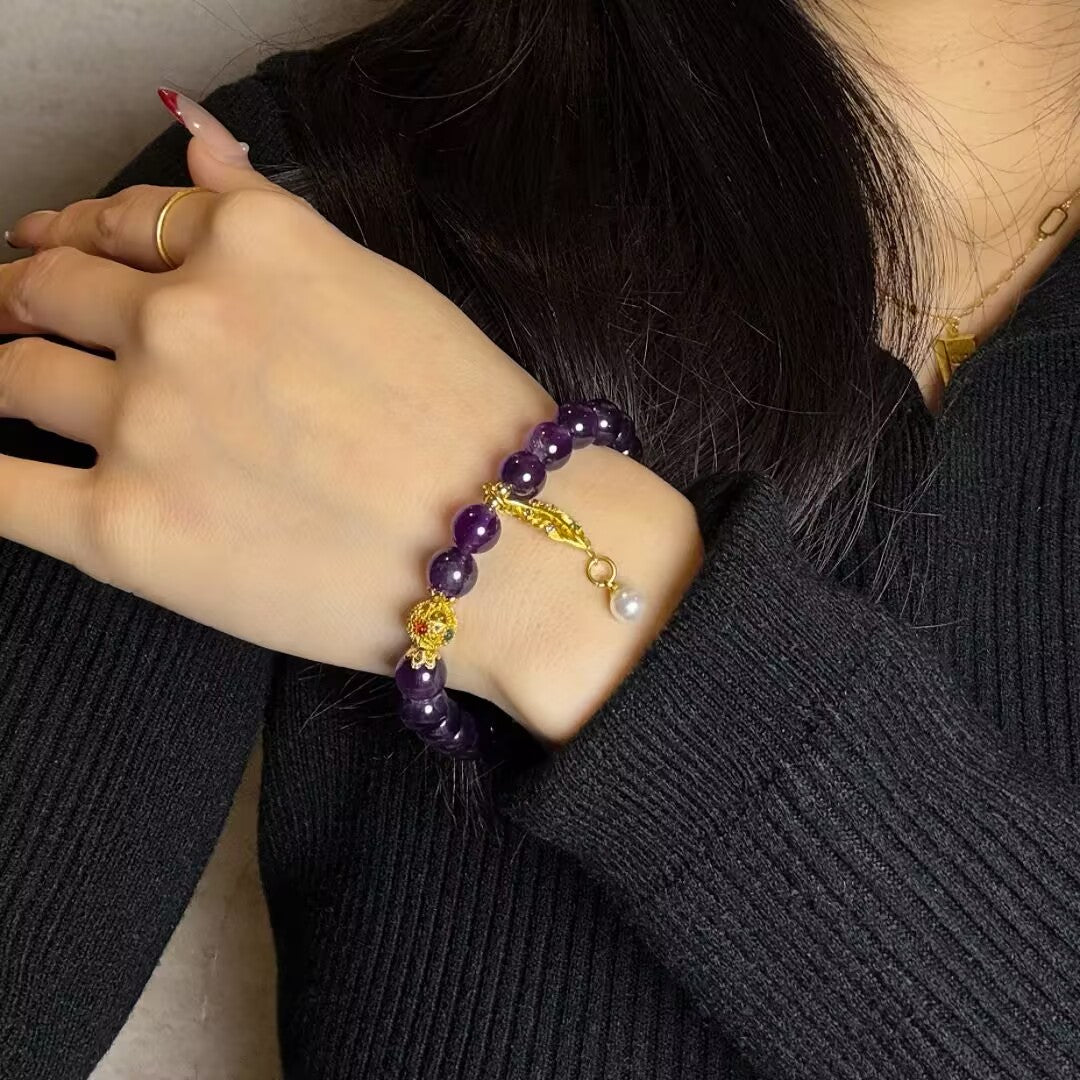 Bracelet en pierres d'améthyste facettées, orné d'une perle blanche suspendue à un fermoir feuille doré et d'une petite pièce dorée ornée de zircons rouges sur le poignet d'une femme.