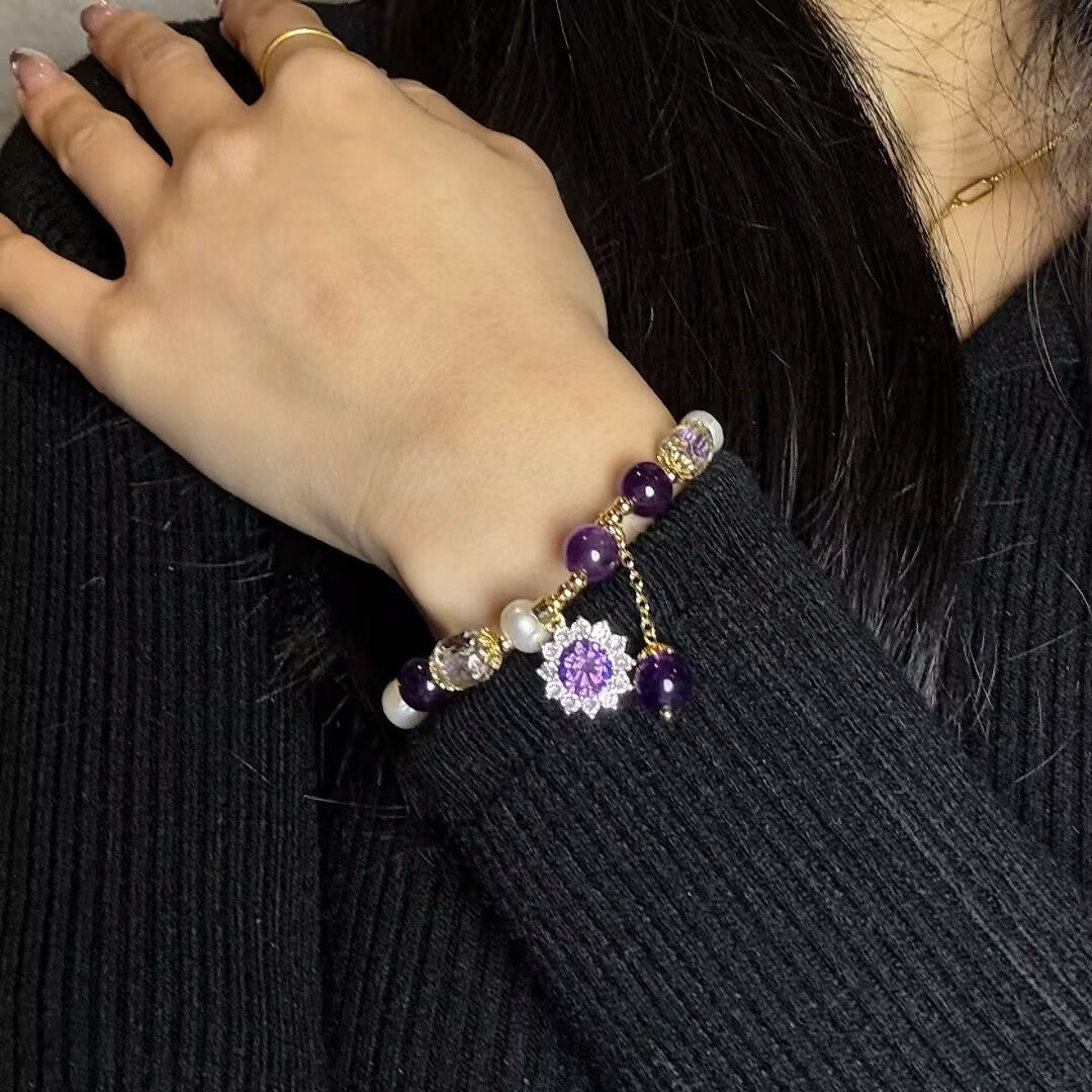 Bracelet artisanal avec des perles d'améthyste, des perles blanches nacrées, des cristaux transparents et un pendentif en forme de fleur violette ornée de strass, porté au poignet.