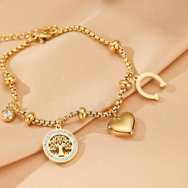 Bracelet en or avec breloques d'un arbre de vie scintillant, d'un cœur lisse, d'un fer à cheval ouvert et de perles sur fond de tissu beige.