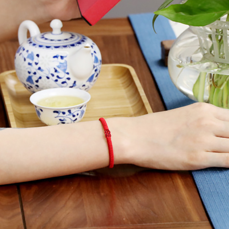 Bracelet en fil rouge avec un nœud double huit stylisé, porté au poignet d'une personne, avec une théière et une tasse en porcelaine à motifs bleus en arrière-plan.