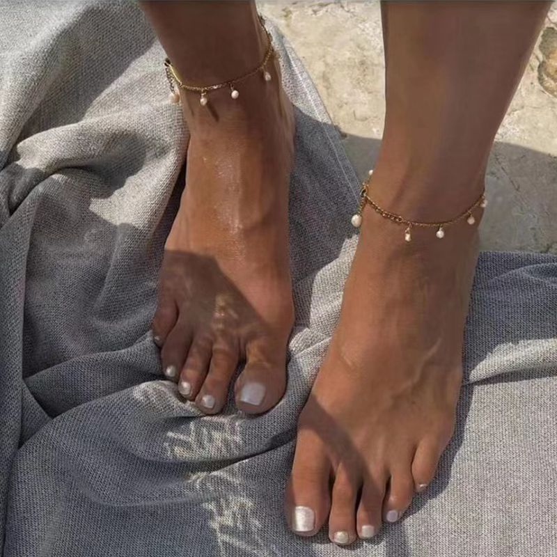 Bracelet de cheville plaqué or avec de petites perles, porté par une femme aux pieds bronzés aux ongles peints en blanc, posés sur une serviette grise.
