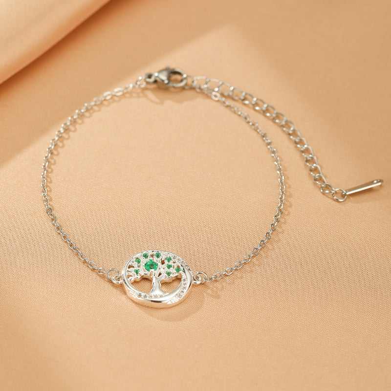 Bracelet avec pendentif arbre de vie en argent serti de petites pierres émeraude et de cristaux brillants sur fond beige.