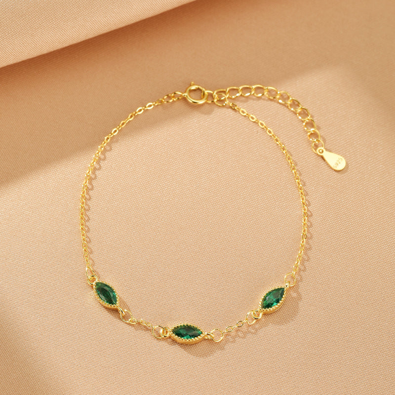 Bracelet fin en argent plaqué or avec trois pierres émeraude en forme de navette, sur fond beige satiné