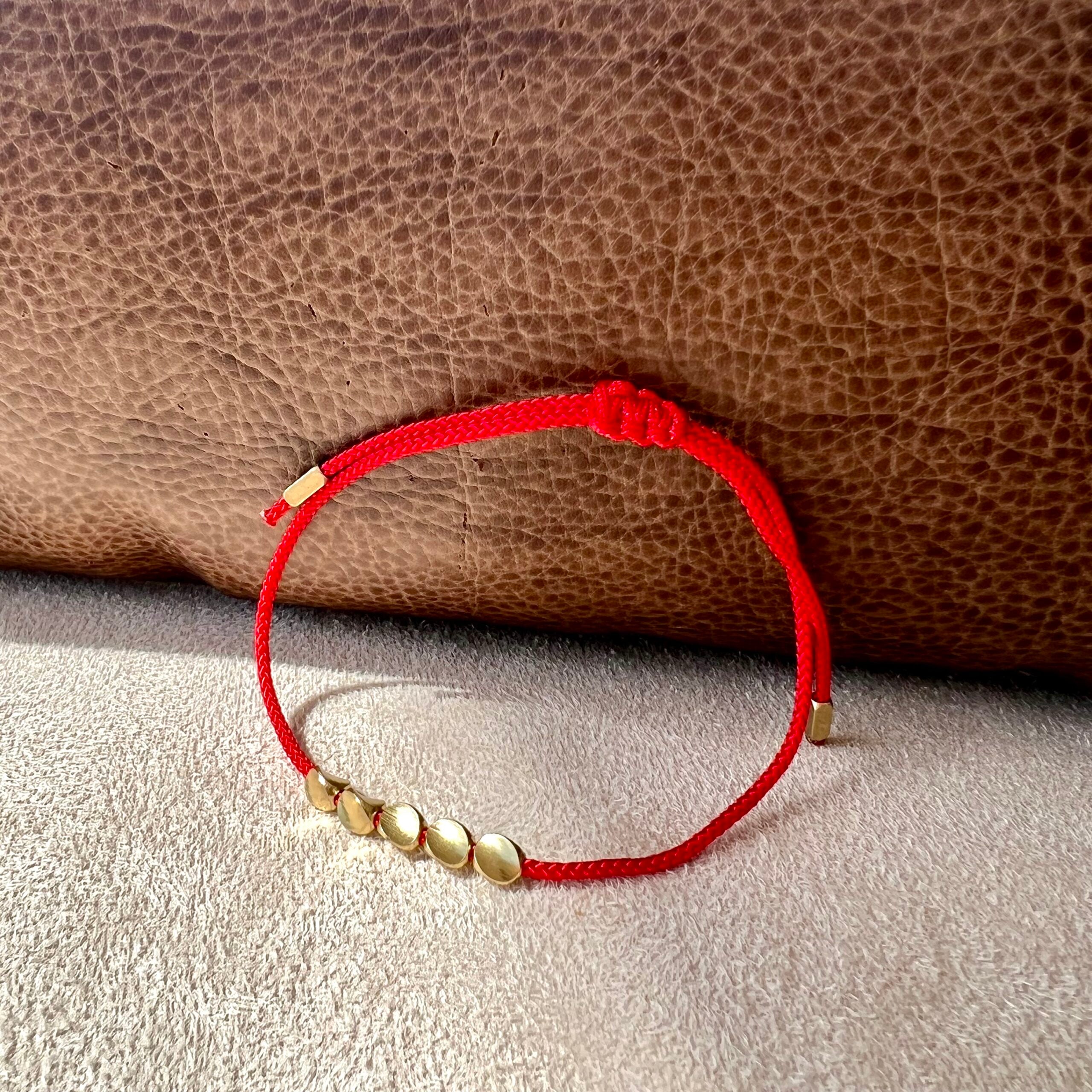 Bracelet fil rouge avec cinq pampilles dorées disposées en arc de cercle, sur un fond texturé marron et gris.