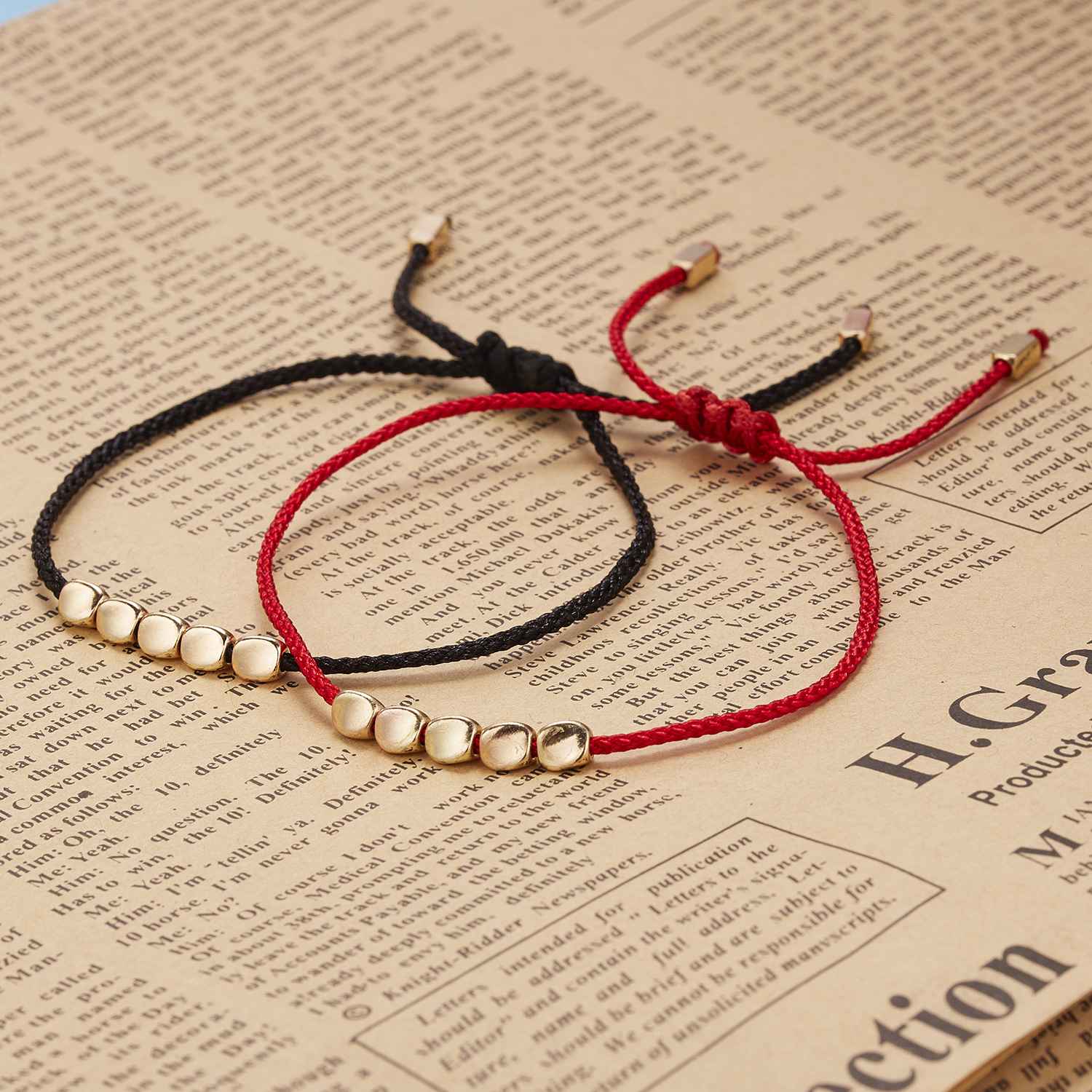 Bracelet Fil Rouge "Lumière et Protection" en fil rouge et noir avec des perles dorées, posé sur un journal.