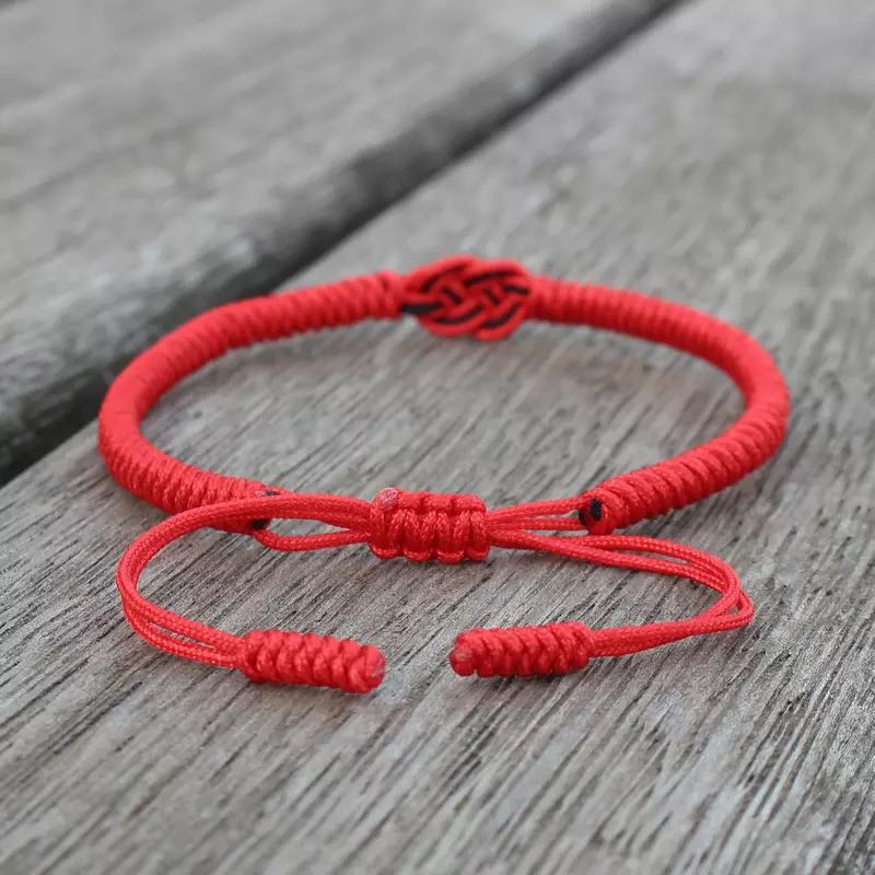 Bracelet fil rouge tibétain avec un nœud central complexe, fermeture coulissante réglable et bouts tressés, posé sur un fond en bois vieilli.